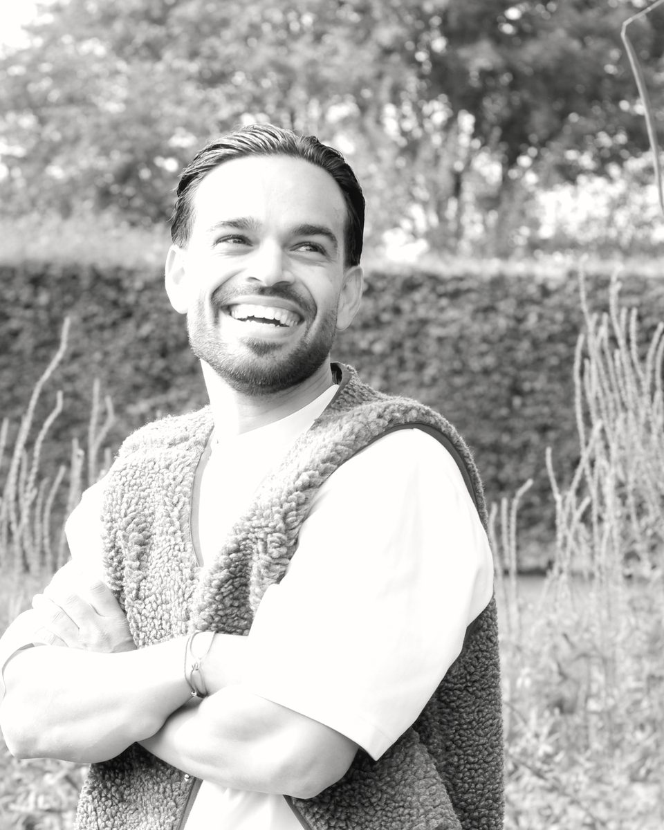 Zwart-witfoto van Alex van Overdijk met de armen over elkaar, lachend in een teddy bodywarmer